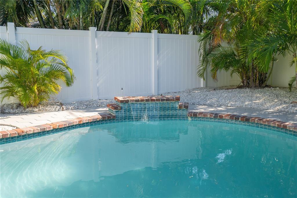 A swimming pool with a brick-trimmed water feature sits in front of a white privacy fence — 3310 E MARITANA DRIVE, ST PETE BEACH, FL