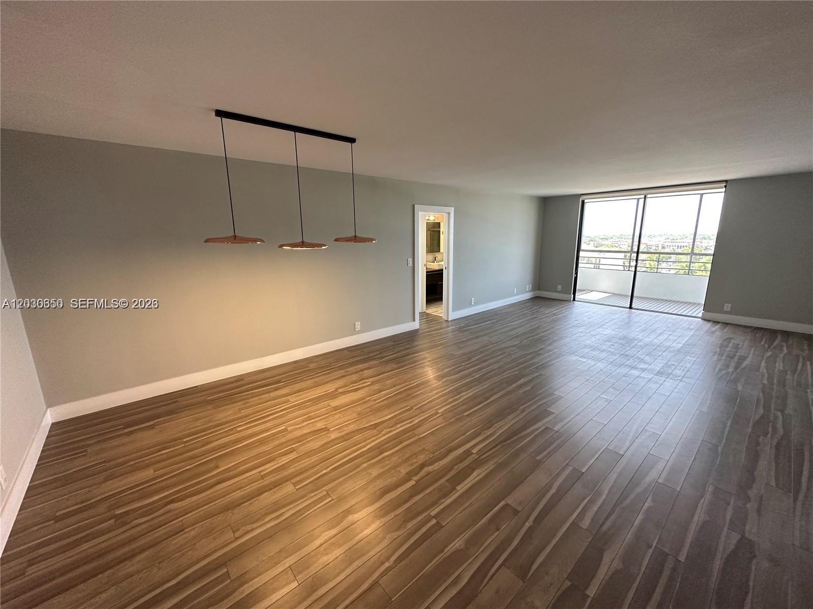 An empty room features wood-look flooring, a three-light pendant fixture, and a sliding glass door — 600 Three Islands Blvd # 705, Hallandale Beach FL 33009, HALLANDALE BEACH, FL