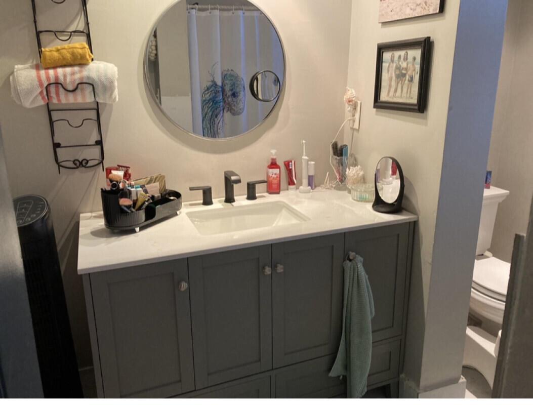 A bathroom vanity features a grey cabinet, white countertop, rectangular sink, and a round mirror — 3550 Blue Lake Drive # 404, Pompano Beach FL 33064, POMPANO BEACH, FL