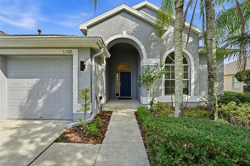 The exterior of a gray house features a single-car garage, an arched entryway, and landscaping — 12018 NORTHUMBERLAND DRIVE, TAMPA, FL