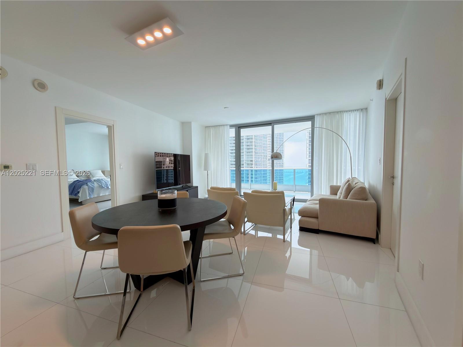 A modern living and dining area features white tile floors, neutral furniture, and a balcony view — 200 Biscayne Boulevard Way # 4104, Miami FL 33131, MIAMI, FL