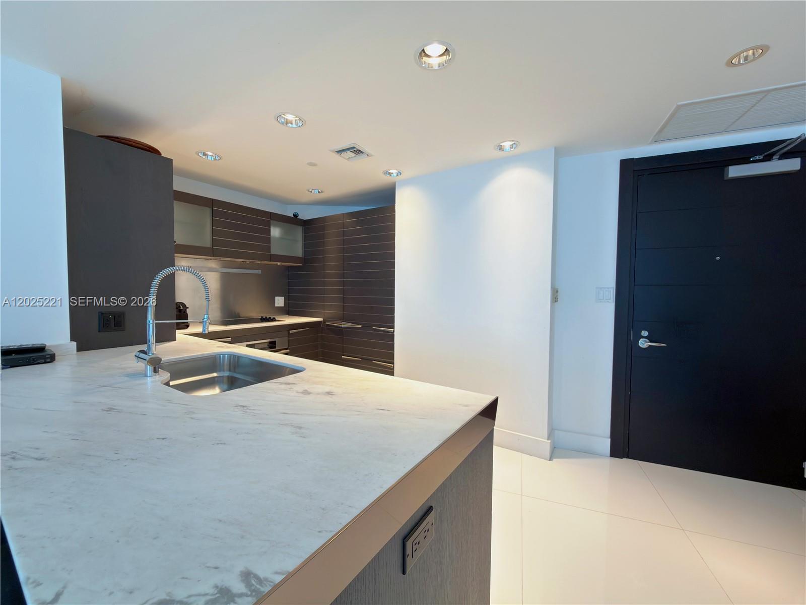 A modern kitchen features a marble-topped island, dark cabinetry, and white tile flooring — 200 Biscayne Boulevard Way # 4104, Miami FL 33131, MIAMI, FL