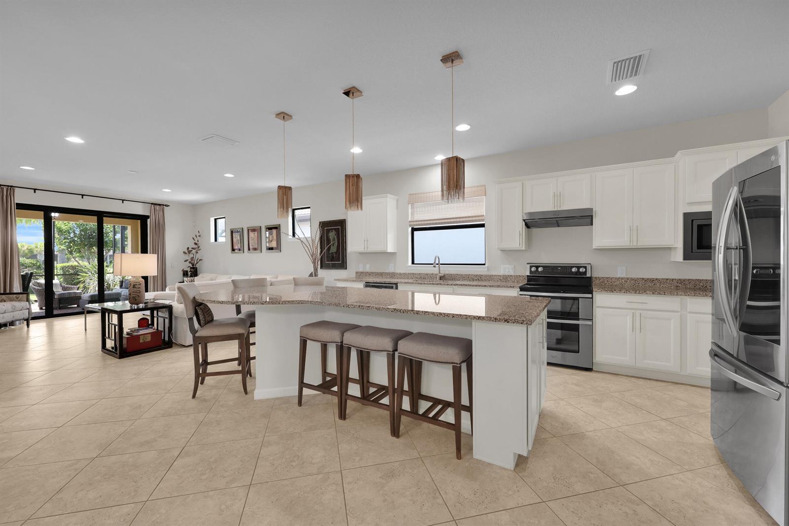 An open-concept kitchen with a granite island, white cabinets, and tile flooring overlooks a living area — 5040 Pendelton Square, Vero Beach FL 32967, VERO BEACH, FL