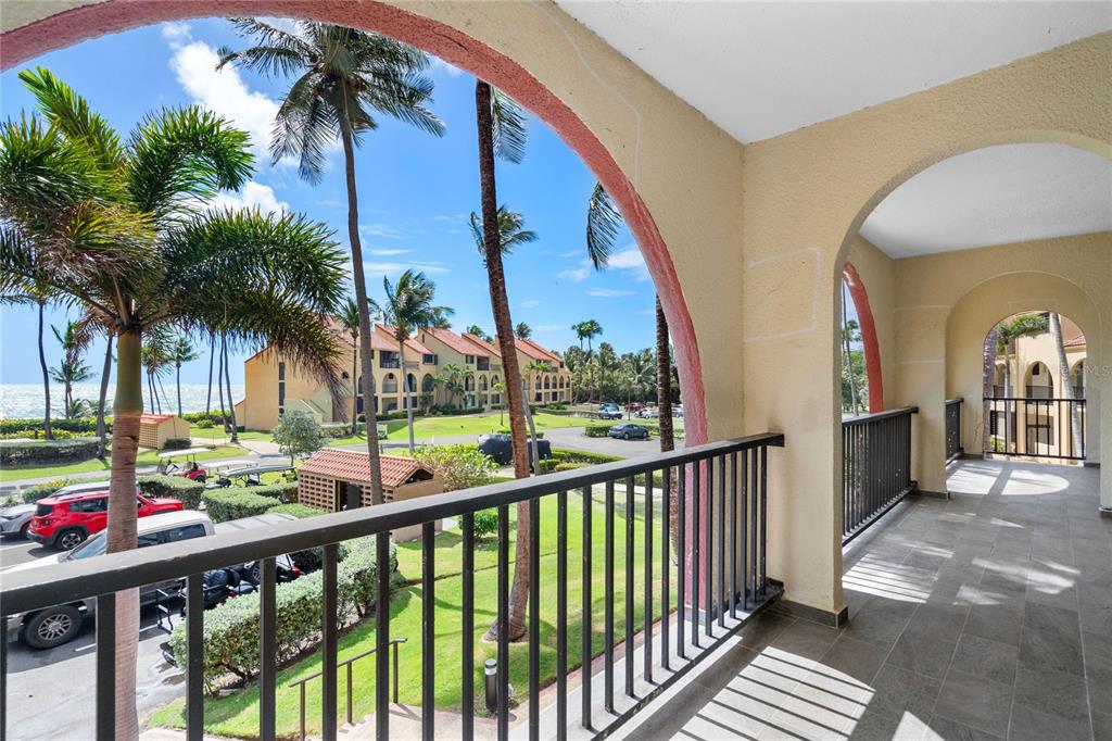 A tiled balcony with arched openings overlooks a parking lot, palm trees, and the ocean — . BEACH VILLAGE UNIT 128, HUMACAO, PR