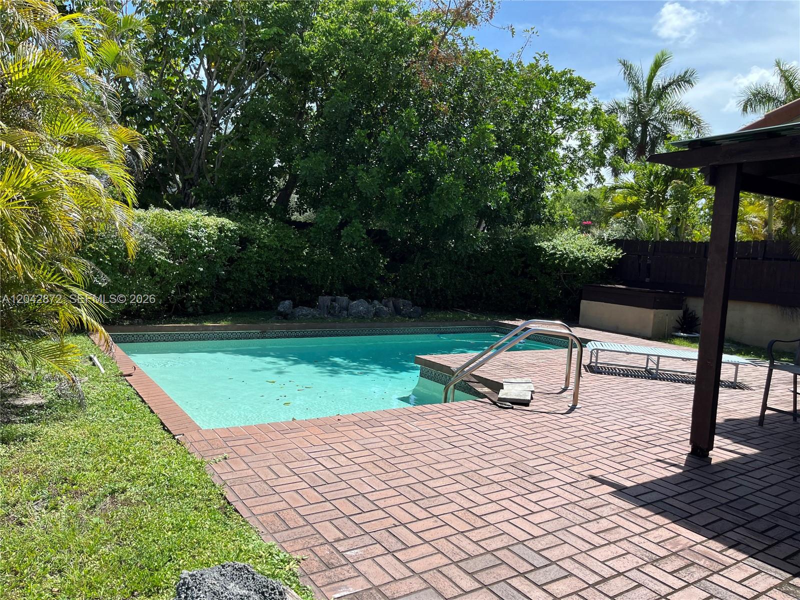 A rectangular swimming pool sits in a backyard with a brick patio and lush landscaping — 1020 NE 202nd Ter, Miami FL 33179, MIAMI, FL