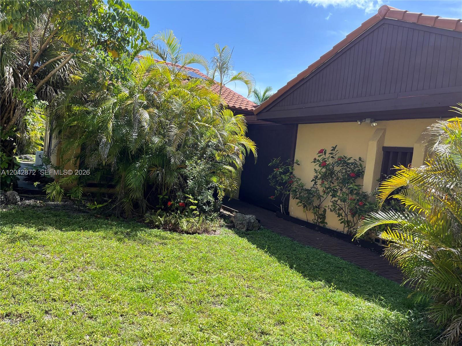 A yellow house exterior features a brown roof, a brick walkway, and lush tropical landscaping — 1020 NE 202nd Ter, Miami FL 33179, MIAMI, FL
