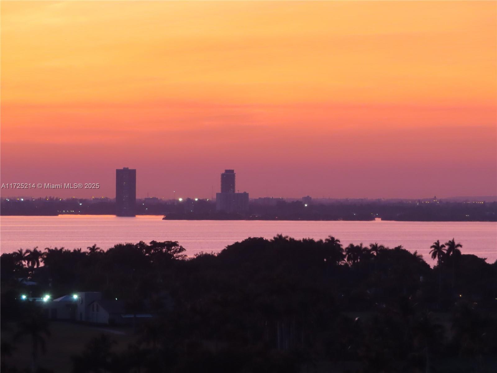 A view of a sunset over a body of water with distant buildings and trees — 5701 Collins Ave # 1011, Miami Beach FL 33140, MIAMI BEACH, FL