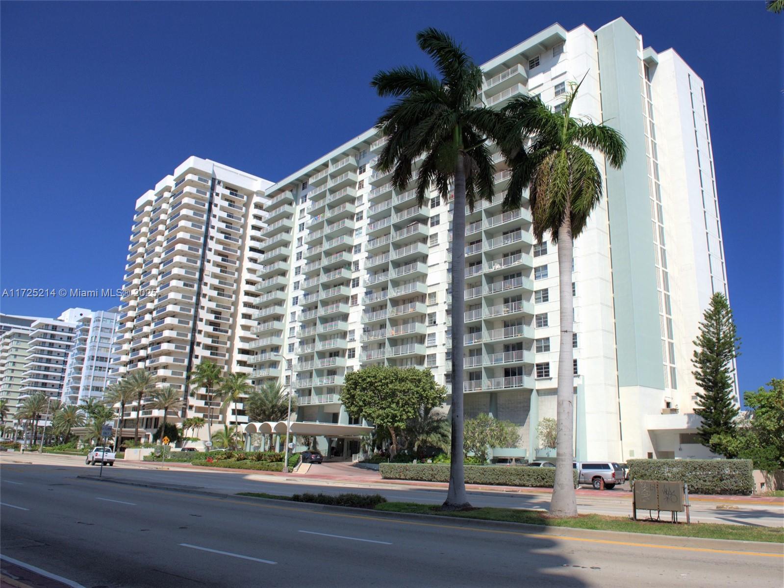 A multi-story residential building with balconies stands behind palm trees and a paved road — 5701 Collins Ave # 1011, Miami Beach FL 33140, MIAMI BEACH, FL
