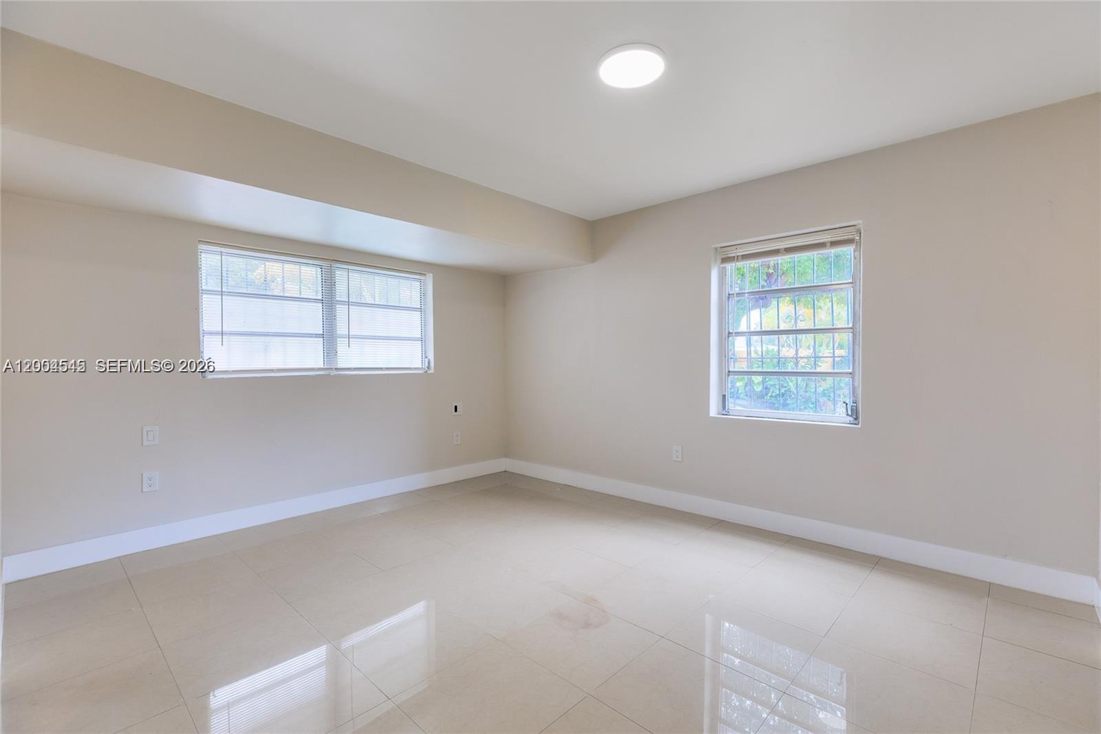 An empty room features beige walls, light-colored tile flooring, two windows, and a ceiling light — 14601 NW 10th Ave # 1, Miami FL 33168, MIAMI, FL