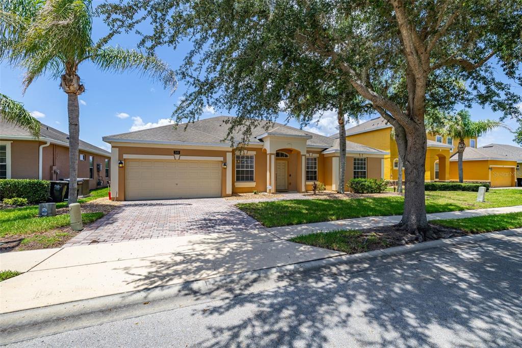 A single-story tan house with a two-car garage and a paved driveway under large trees — 349 YELLOW SNAPDRAGON DRIVE, DAVENPORT, FL