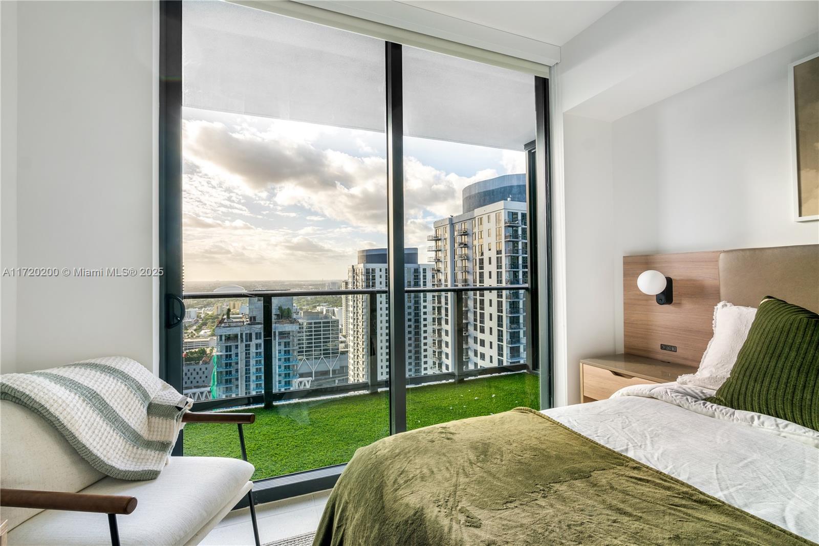 A bedroom features a bed, an armchair, and a balcony with artificial grass overlooking city buildings — 601 NE 1 AVE # 3604, Miami FL 33132, MIAMI, FL
