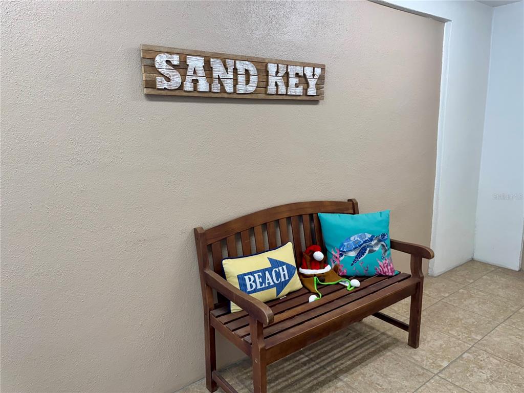 A wooden bench with two decorative pillows sits against a beige wall under a sign — 1230 GULF BOULEVARD UNIT 1402, CLEARWATER BEACH, FL