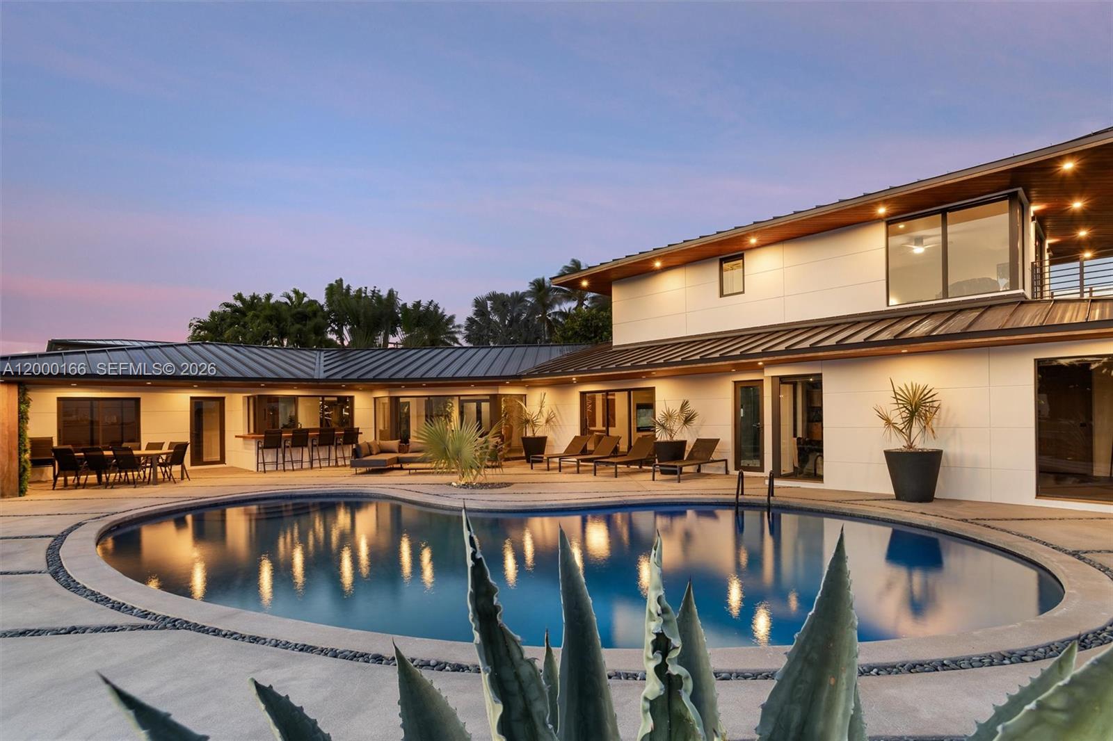 A modern two-story house with a metal roof overlooks a curved swimming pool at dusk — 1982 NE 119th Rd, North Miami FL 33181, NORTH MIAMI, FL