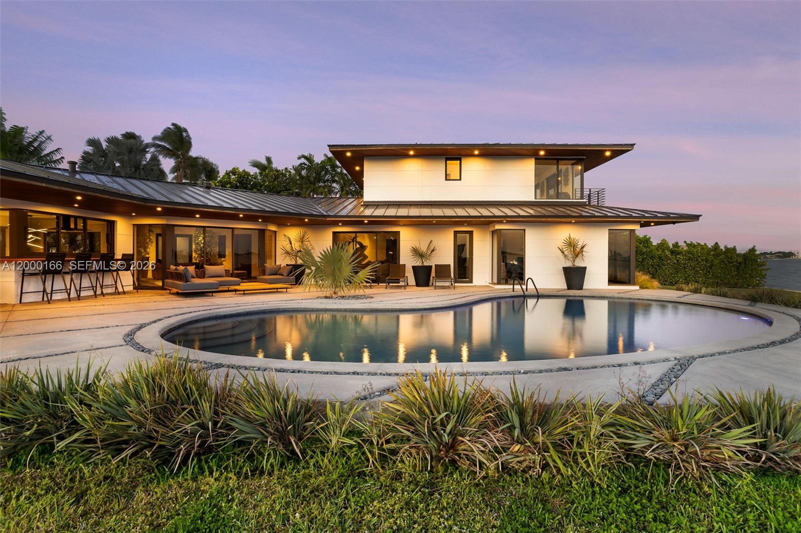 A modern two-story house features a curved swimming pool and a patio at sunset — 1982 NE 119th Rd, North Miami FL 33181, NORTH MIAMI, FL