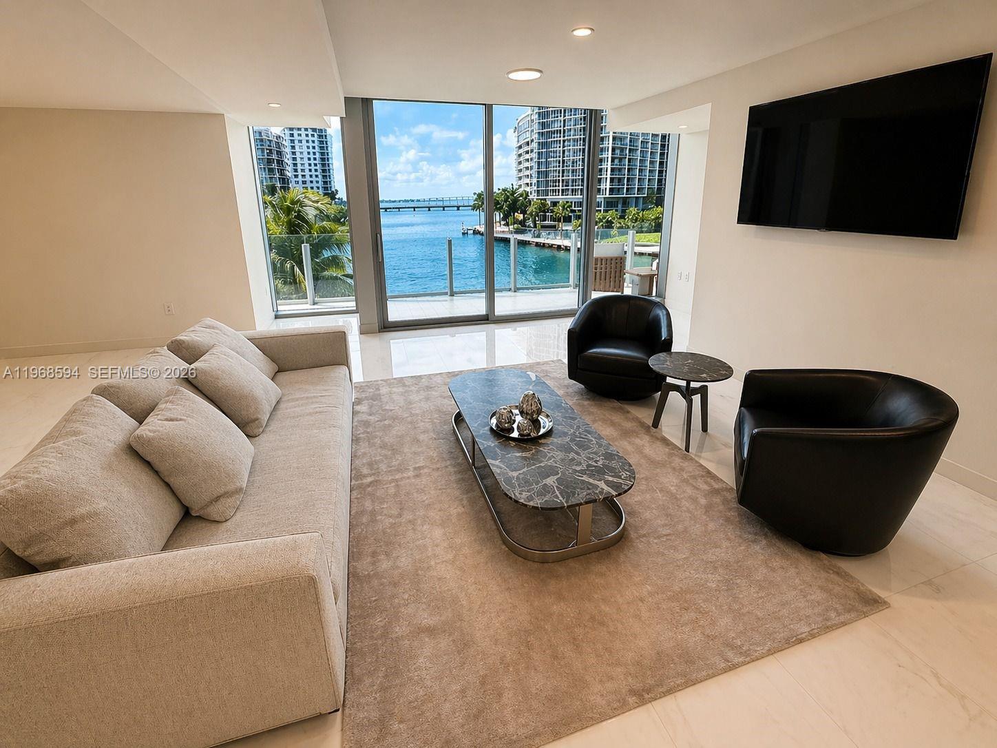 A living room features a beige sofa, two black armchairs, and a waterfront balcony view — 300 Biscayne Blvd Way # 303, Miami FL 33131, MIAMI, FL
