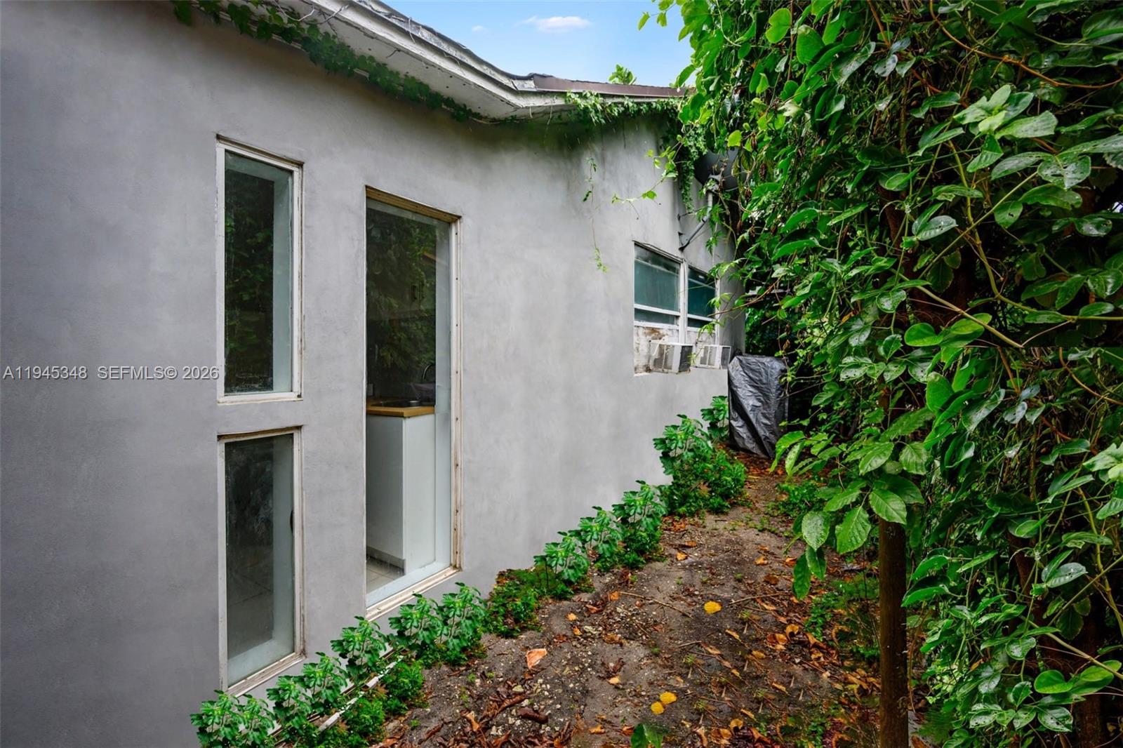 The exterior of a gray stucco house features tall windows and overgrown vegetation — 348 NE 58th St, Miami FL 33137, MIAMI, FL