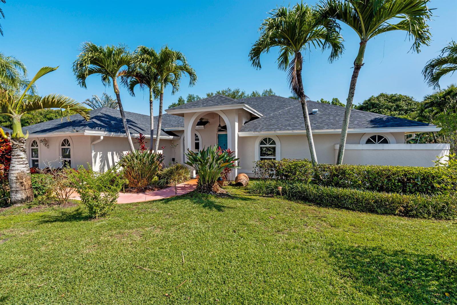 A single-story house with a light-colored exterior, arched entryway, and palm trees in the yard — 15495 Rolling Meadows Circle, Wellington FL 33414, WELLINGTON, FL