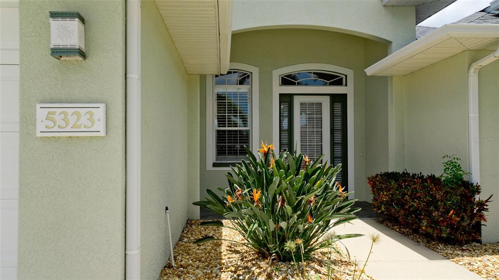 The exterior entrance of a house features a light green stucco facade and a bird-of-paradise plant — 5323 90TH AVENUE CIRCLE E, PARRISH, FL