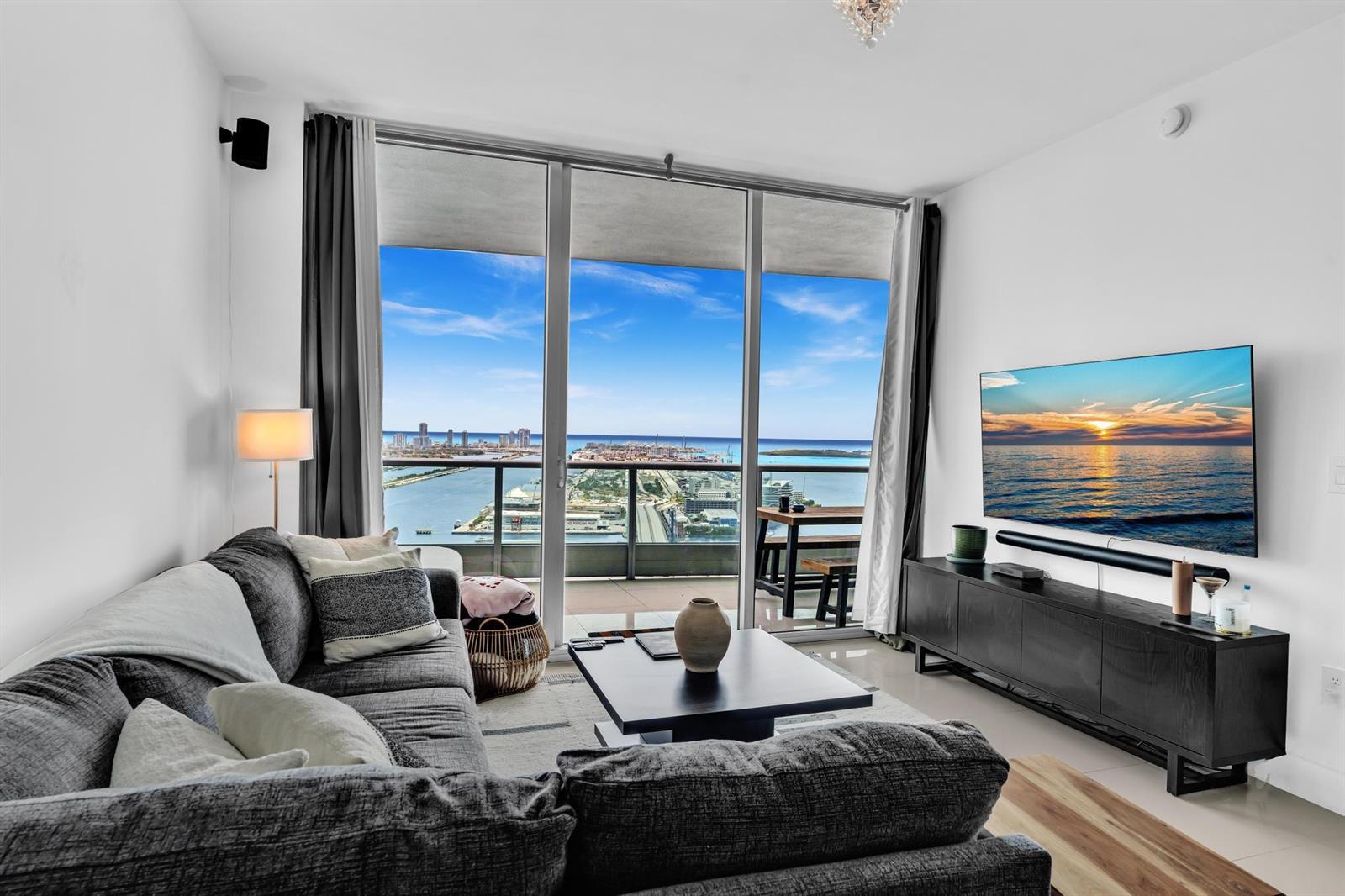 A living room with a gray sectional sofa, television, and balcony overlooking a coastal city — 900 Biscayne Boulevard # 4703, Miami FL 33132, MIAMI, FL