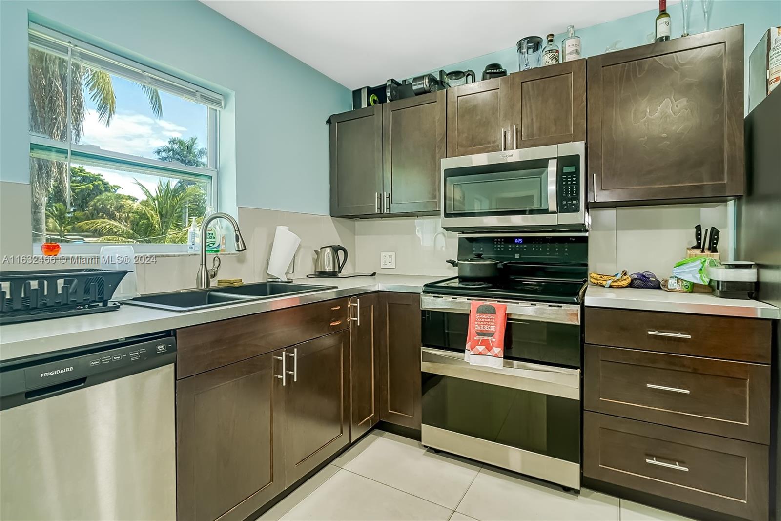 A kitchen features dark wood cabinets, stainless steel appliances, and a window overlooking palm trees — 216 SW 6th St, Hallandale Beach FL 33009, HALLANDALE BEACH, FL