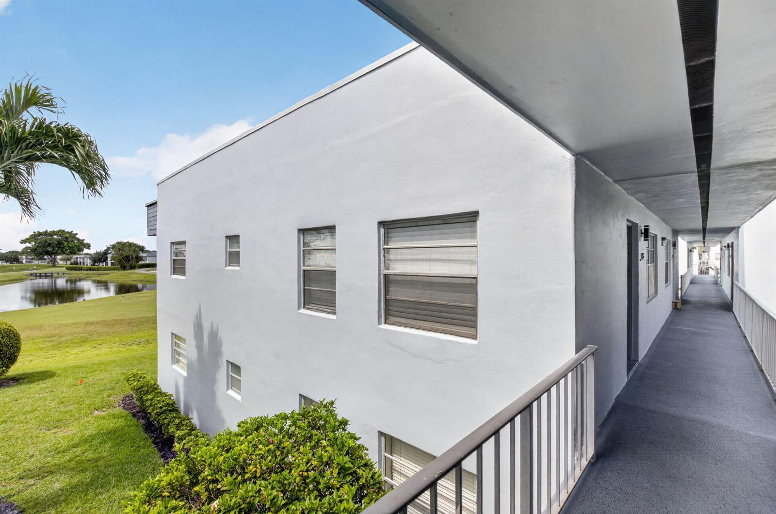An exterior view of a white apartment building with a walkway overlooking a pond — 955 Normandy Trail, Delray Beach FL 33484, DELRAY BEACH, FL