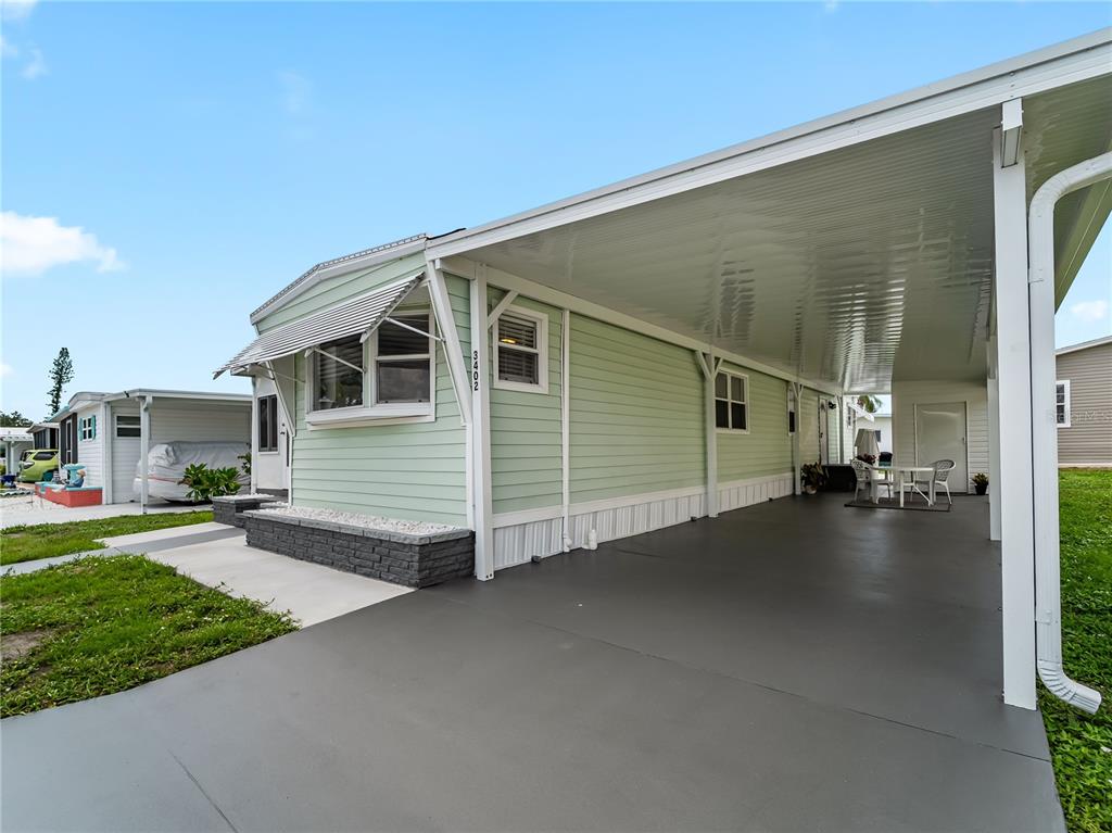 A light green manufactured home features a large attached carport and a brick-accented planter — 3402 SHINGLE OAK TERRACE, SARASOTA, FL