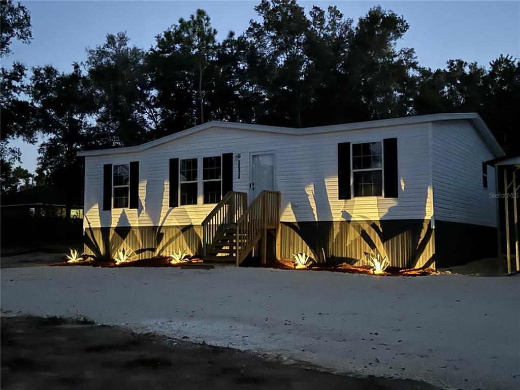 A single-story manufactured home with white siding, dark shutters, and exterior landscape lighting at dusk — 10970 NE 109TH STREET, ARCHER, FL