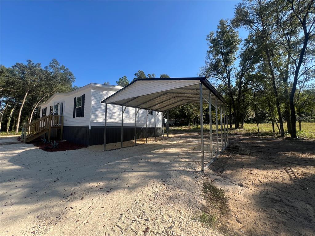 A white mobile home with a metal carport stands on a sandy lot near trees — 10970 NE 109TH STREET, ARCHER, FL