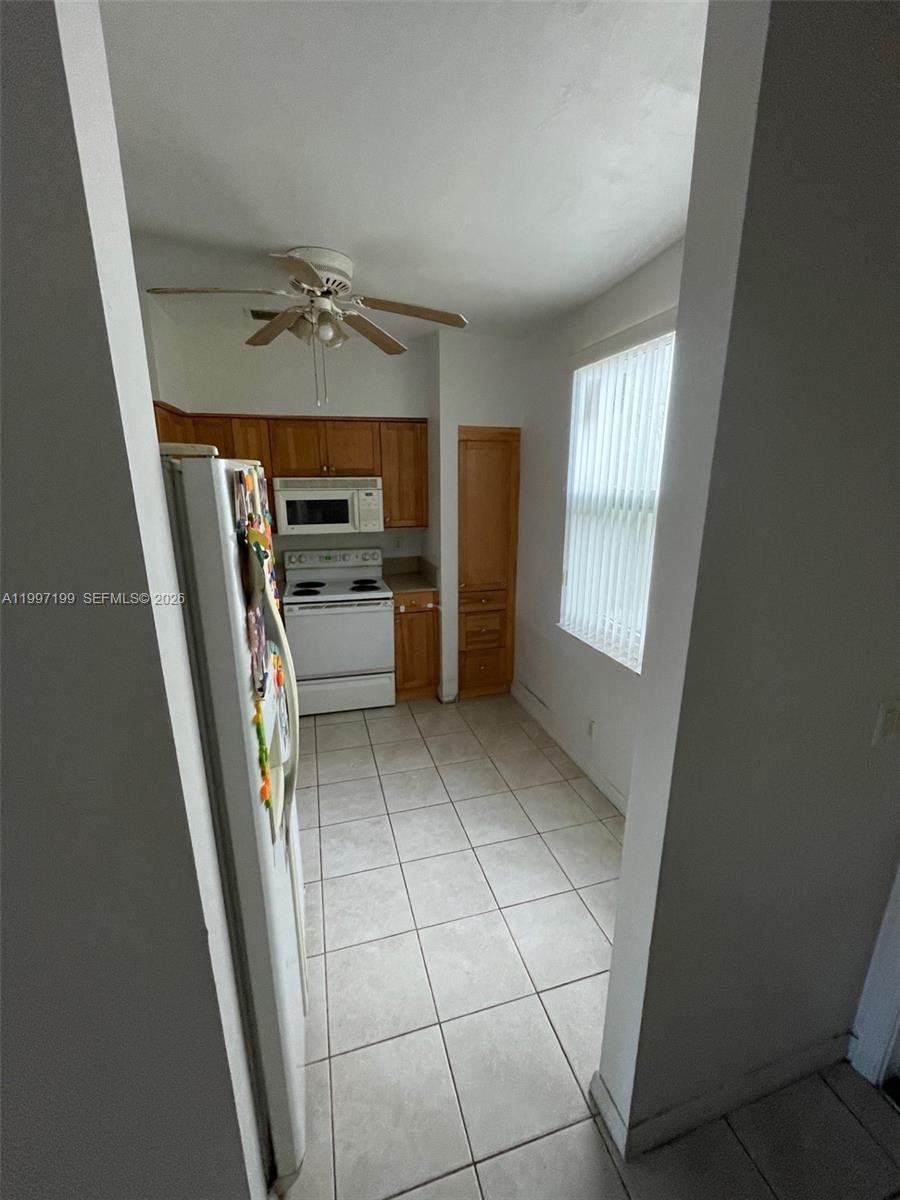 A narrow kitchen features light-colored tile flooring, wooden cabinets, and a white refrigerator — 870 NE 212th Ter # 3, Miami FL 33179, MIAMI, FL