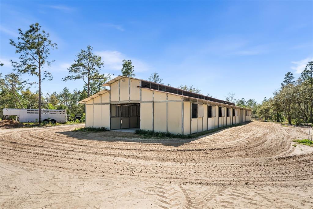 A long, light-colored horse barn with multiple stalls sits on a sandy, cleared lot — 25723 BAREVA ROAD, WEEKI WACHEE, FL