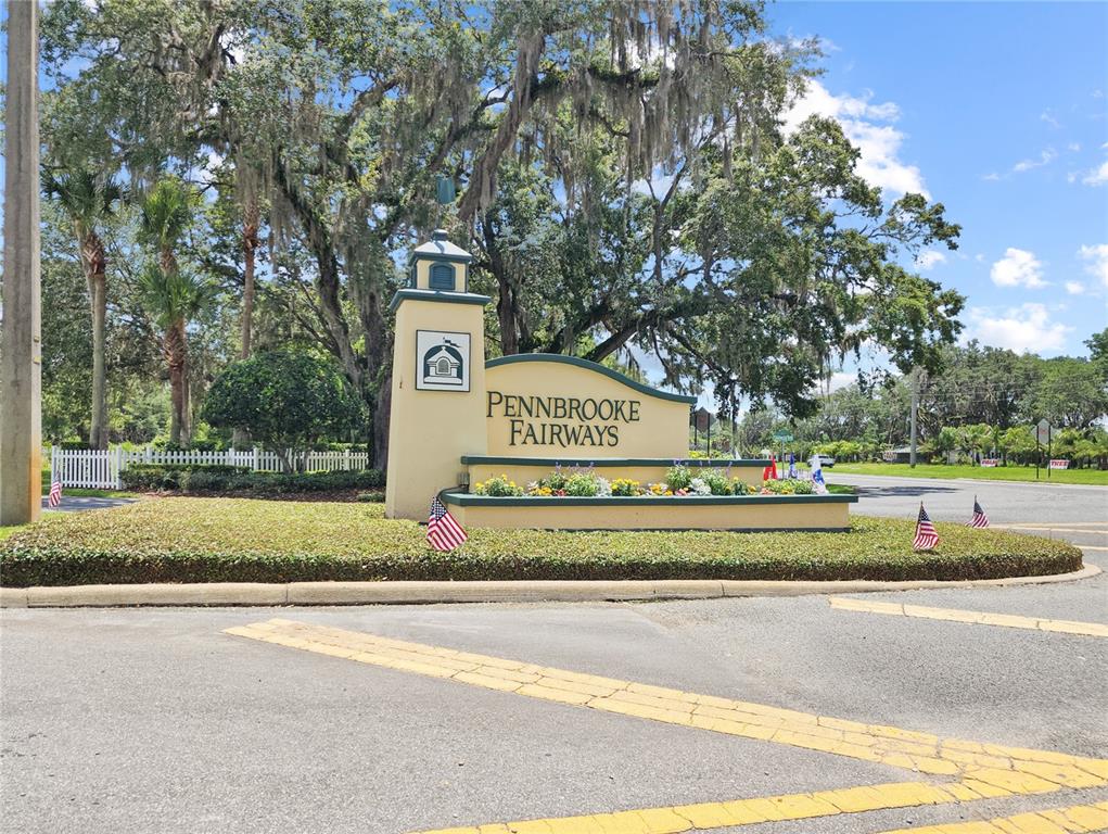 A beige monument sign for Pennbrooke Fairways stands in a landscaped island with American flags — 615 NE COTTAGE PARK LANE, LEESBURG, FL