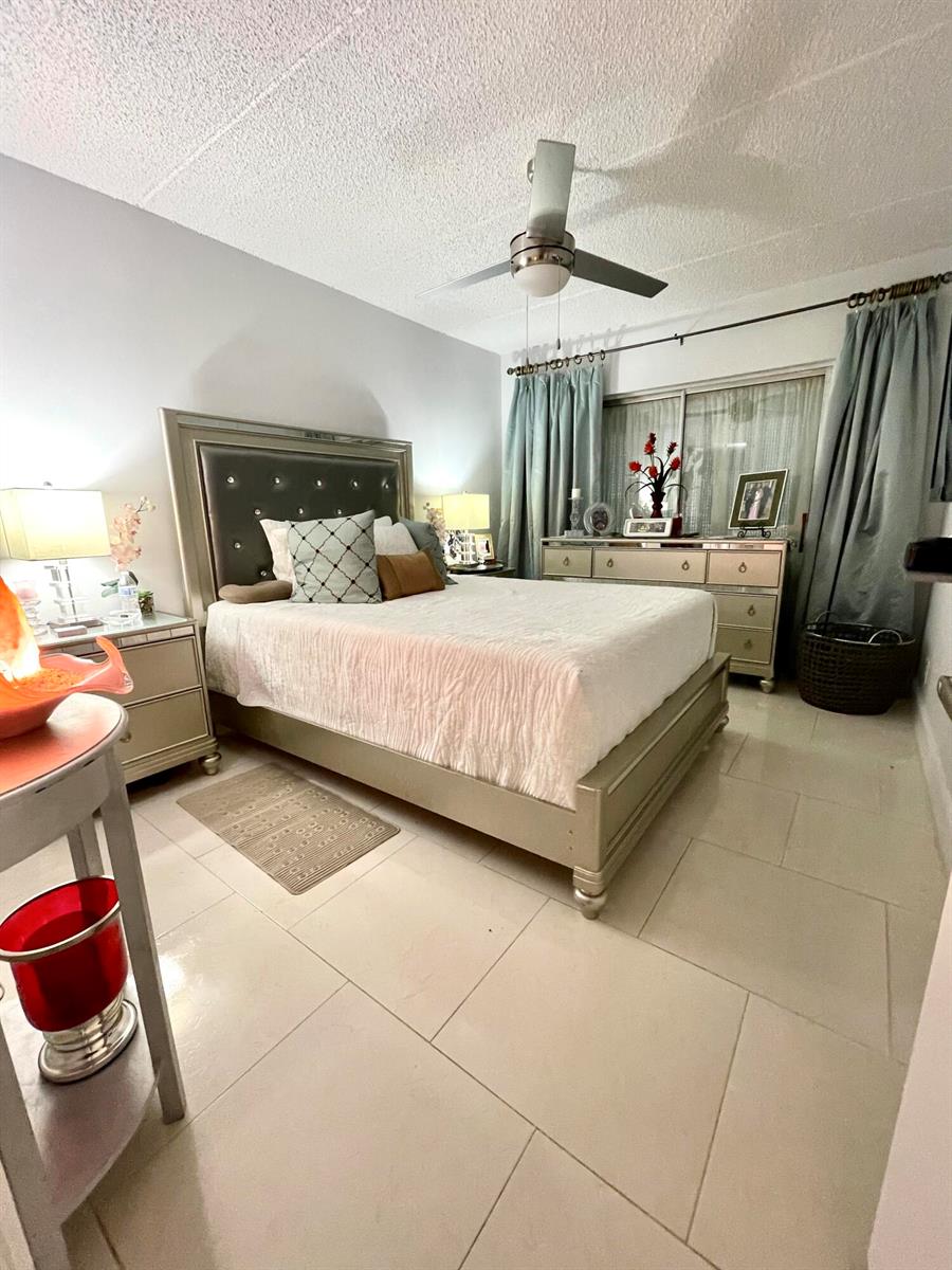 A bedroom features a metallic bed frame, tiled flooring, and light blue curtains — 3200 Springdale Boulevard # 107, Palm Springs FL 33461, PALM SPRINGS, FL