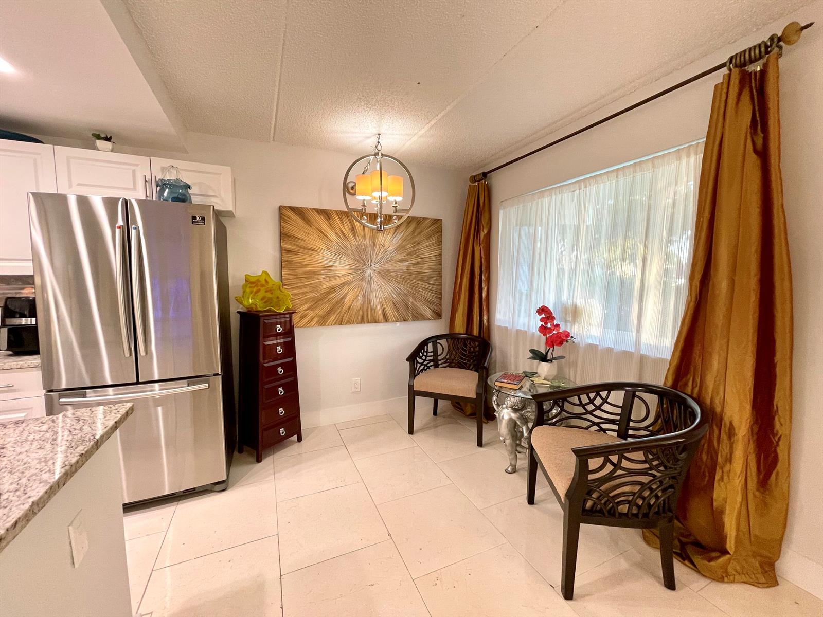 A kitchen dining nook features a stainless steel refrigerator, two dark chairs, and gold curtains — 3200 Springdale Boulevard # 107, Palm Springs FL 33461, PALM SPRINGS, FL