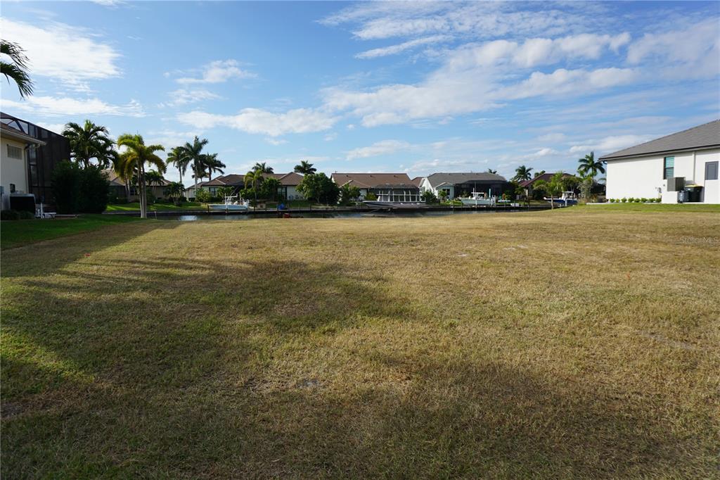 A vacant, grassy waterfront lot sits between two houses under a blue sky — 4019 MALTESE COURT, PUNTA GORDA, FL