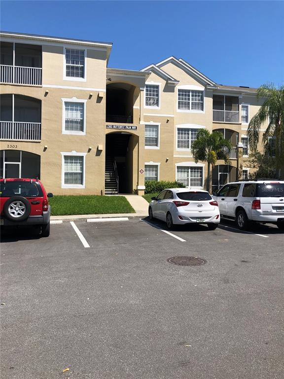 A three-story apartment building with tan stucco siding stands behind a paved parking lot — 8100 PRINCESS PALM LANE UNIT 203, KISSIMMEE, FL