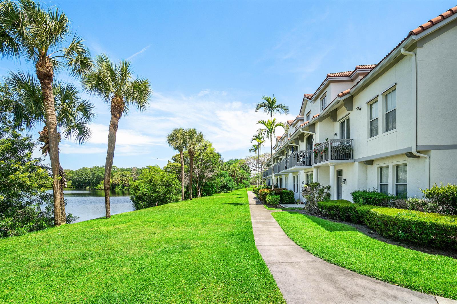 A sidewalk runs alongside a multi-story apartment building with balconies overlooking a lake and lawn — 2052 Alta Meadows Lane Apt 2212, Delray Beach FL 33444, DELRAY BEACH, FL