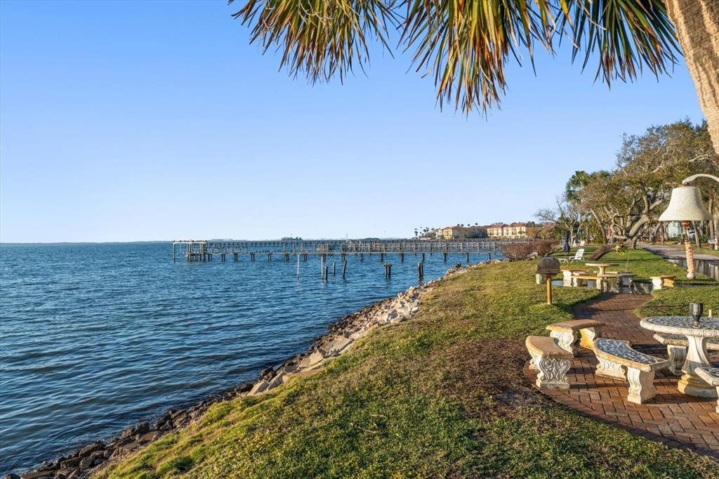 A waterfront patio with stone benches and a table overlooks a pier and the bay — 1101 VICTORIA DRIVE UNIT 7, DUNEDIN, FL