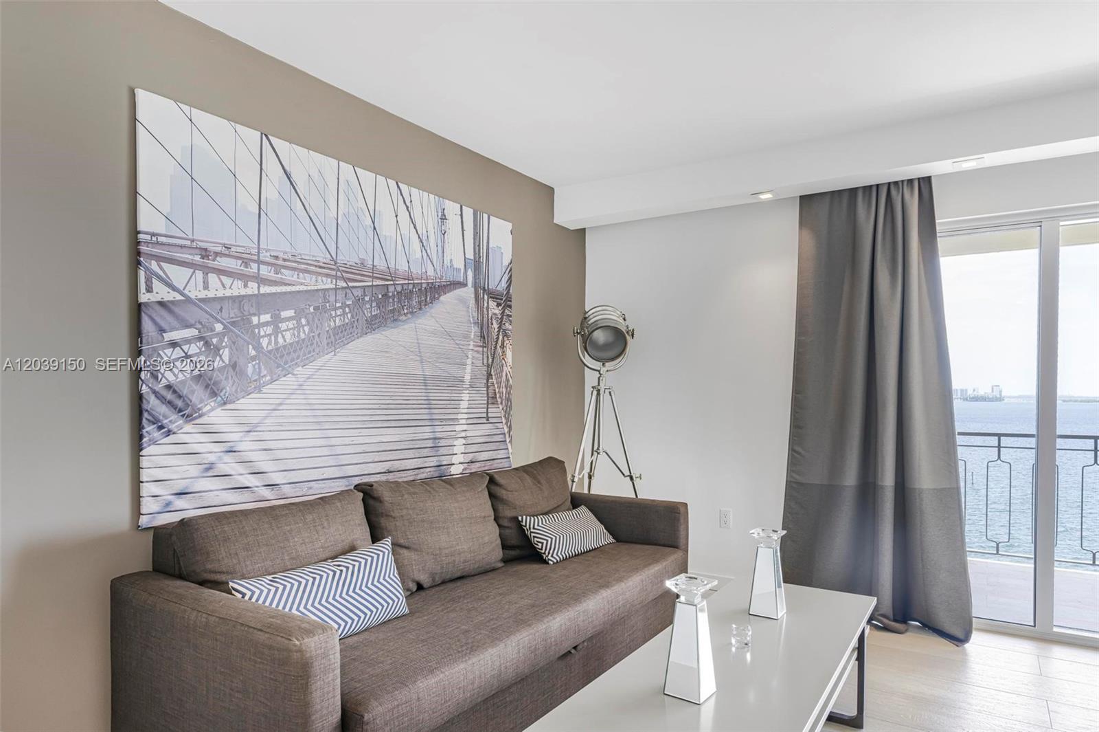 A living room features a large bridge wall art, a brown sofa, and a balcony view — 11111 Biscayne Blvd # 8C, Miami FL 33181, MIAMI, FL