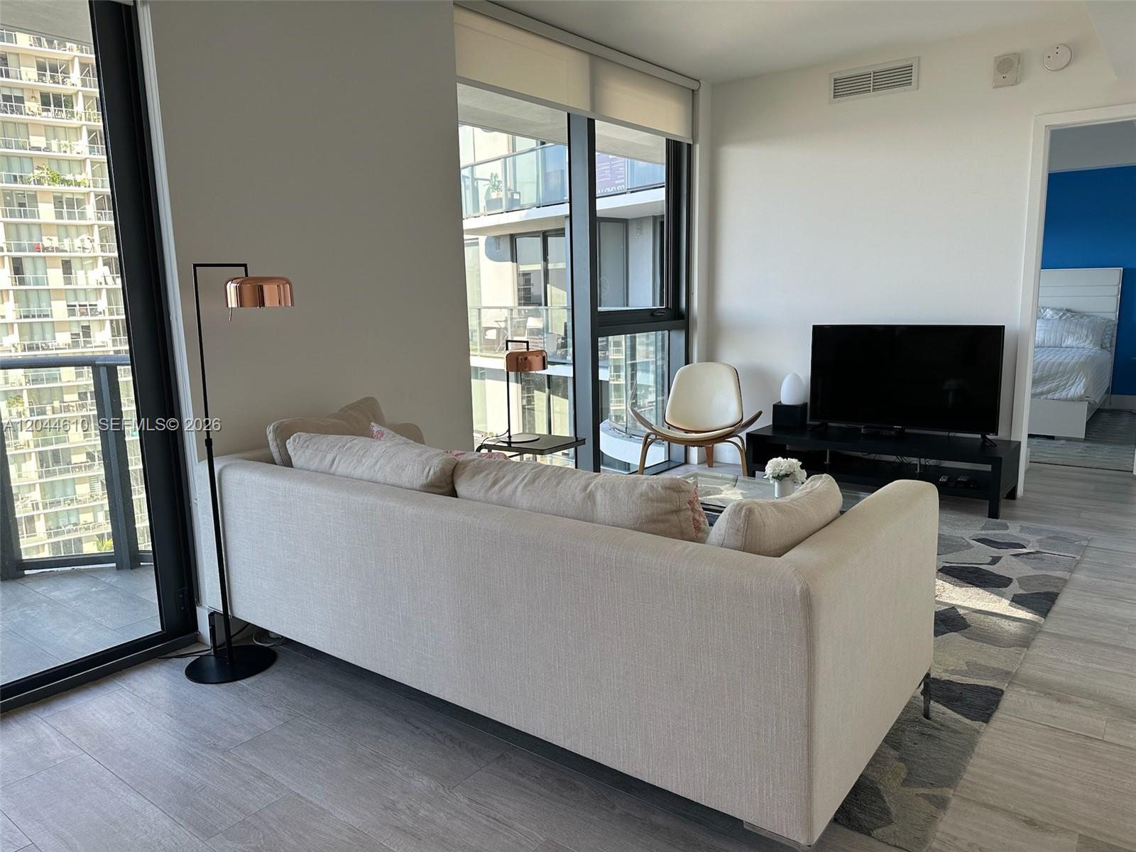 A living room features a beige sofa, a television, and a view of neighboring buildings — 121 NE 34th St # 1915, Miami FL 33137, MIAMI, FL