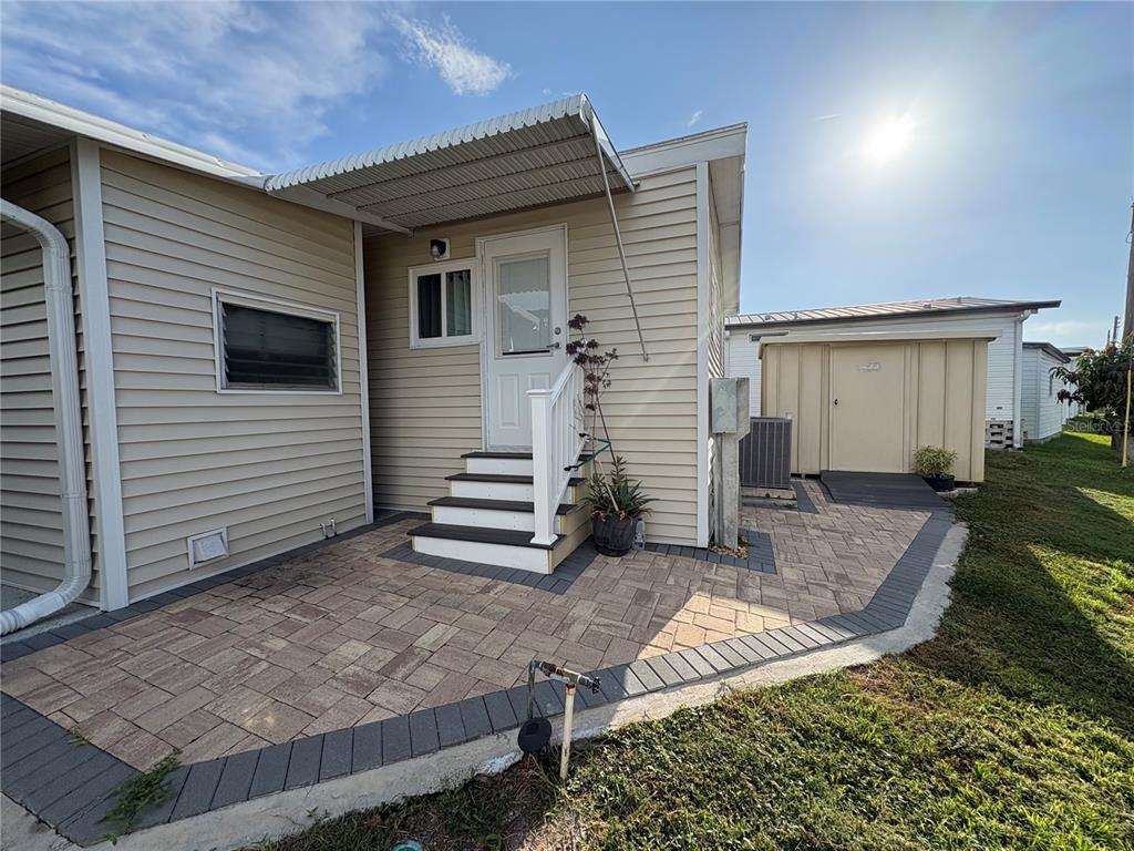 A tan mobile home exterior features a paved patio, white stairs, and a storage shed — 1706 WISCONSIN AVENUE, BRADENTON, FL