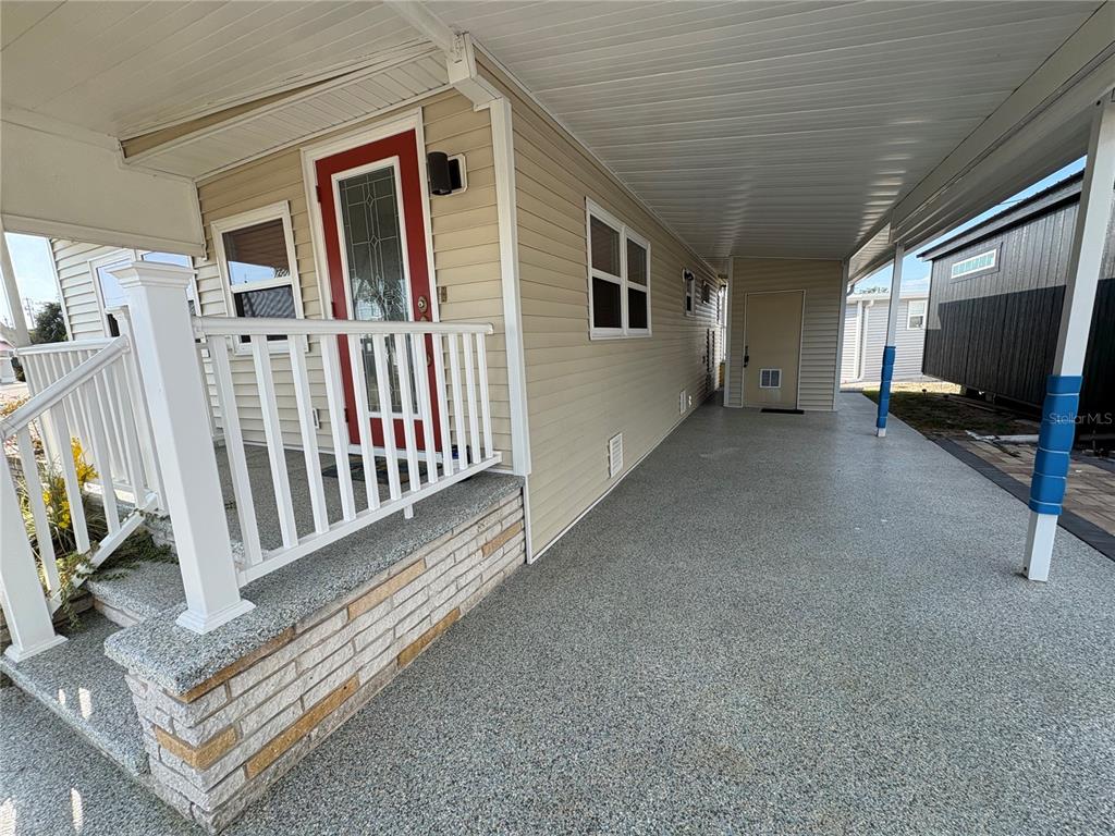 A covered carport with a textured floor leads to a red front door and porch — 1706 WISCONSIN AVENUE, BRADENTON, FL