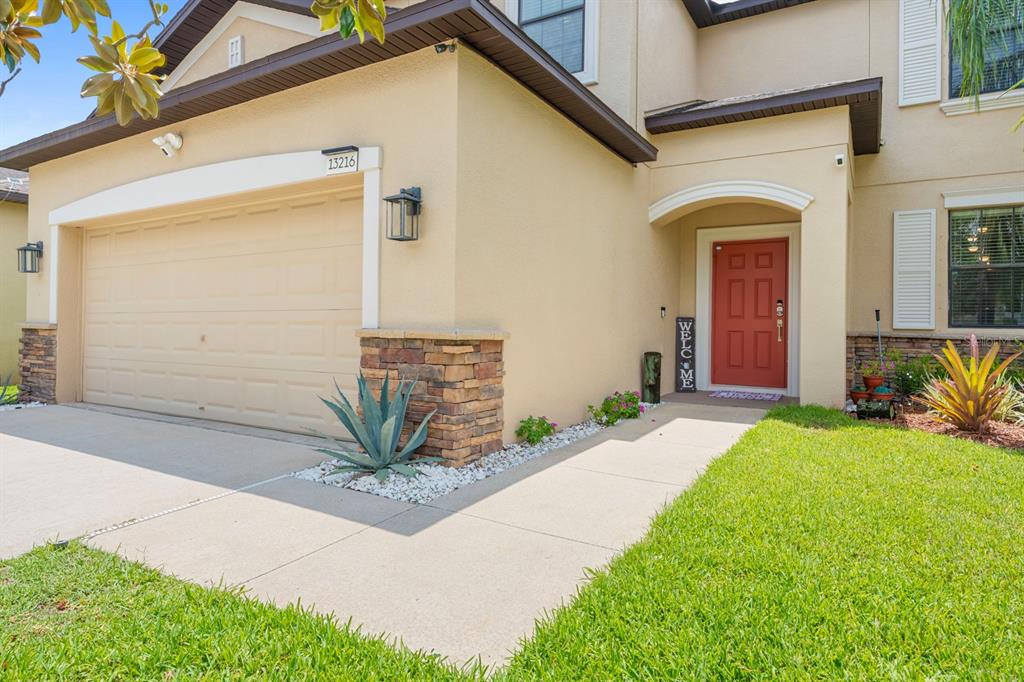 A two-story house exterior features a tan stucco facade, a red front door, and a garage — 13216 WELLINGTON HILLS DRIVE, RIVERVIEW, FL