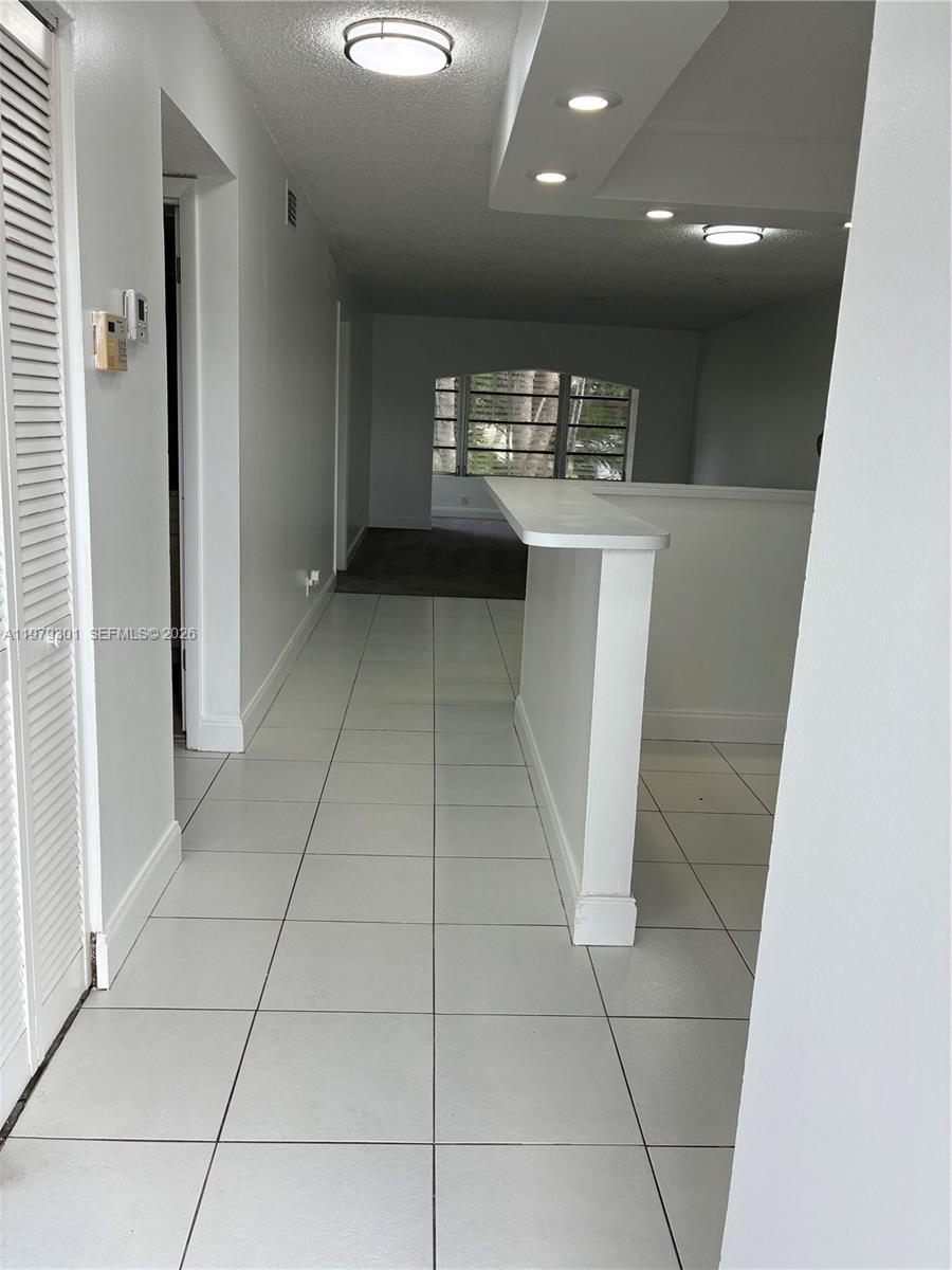 A hallway with white tile flooring leads toward an arched window in a carpeted room — 609 S State Road 7 # 2C, Margate FL 33068, MARGATE, FL