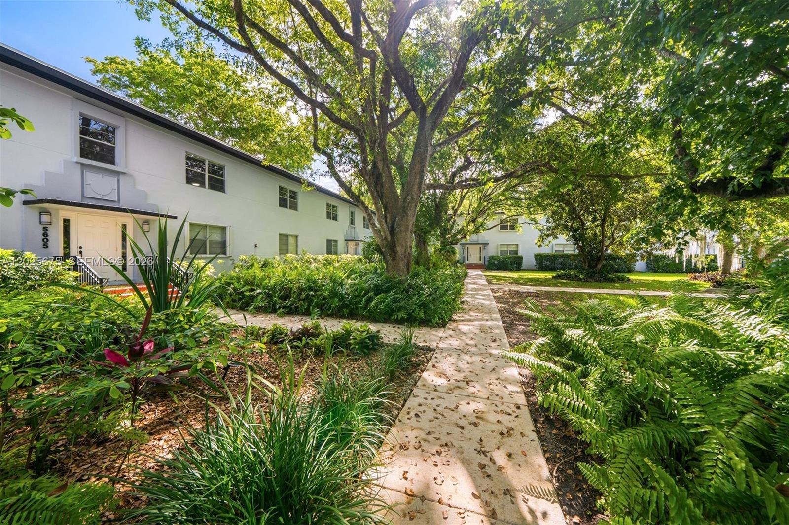 A sidewalk leads through a lush garden toward the entrance of a white apartment building — 5605 SW 80th St # 10D, Miami FL 33143, MIAMI, FL