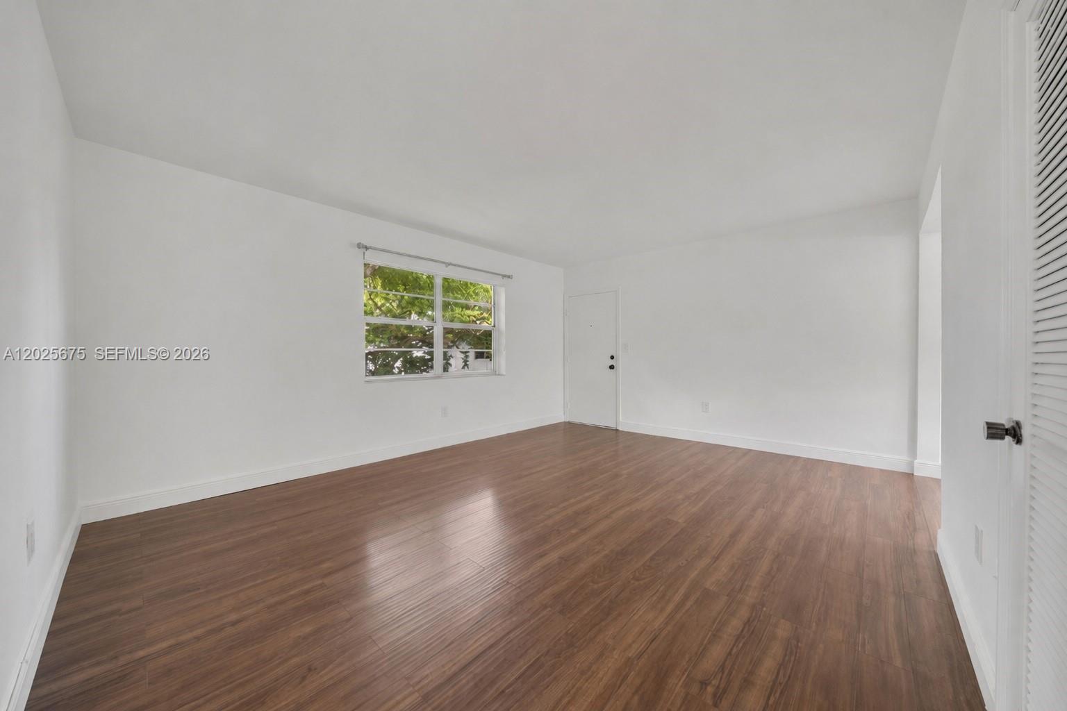 An empty room features white walls, wood-look flooring, a window, and a white door — 5605 SW 80th St # 10D, Miami FL 33143, MIAMI, FL