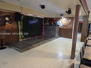 A room features a raised wooden stage with a green wall backdrop and overhead lighting — MIAMI SPRINGS, FL
