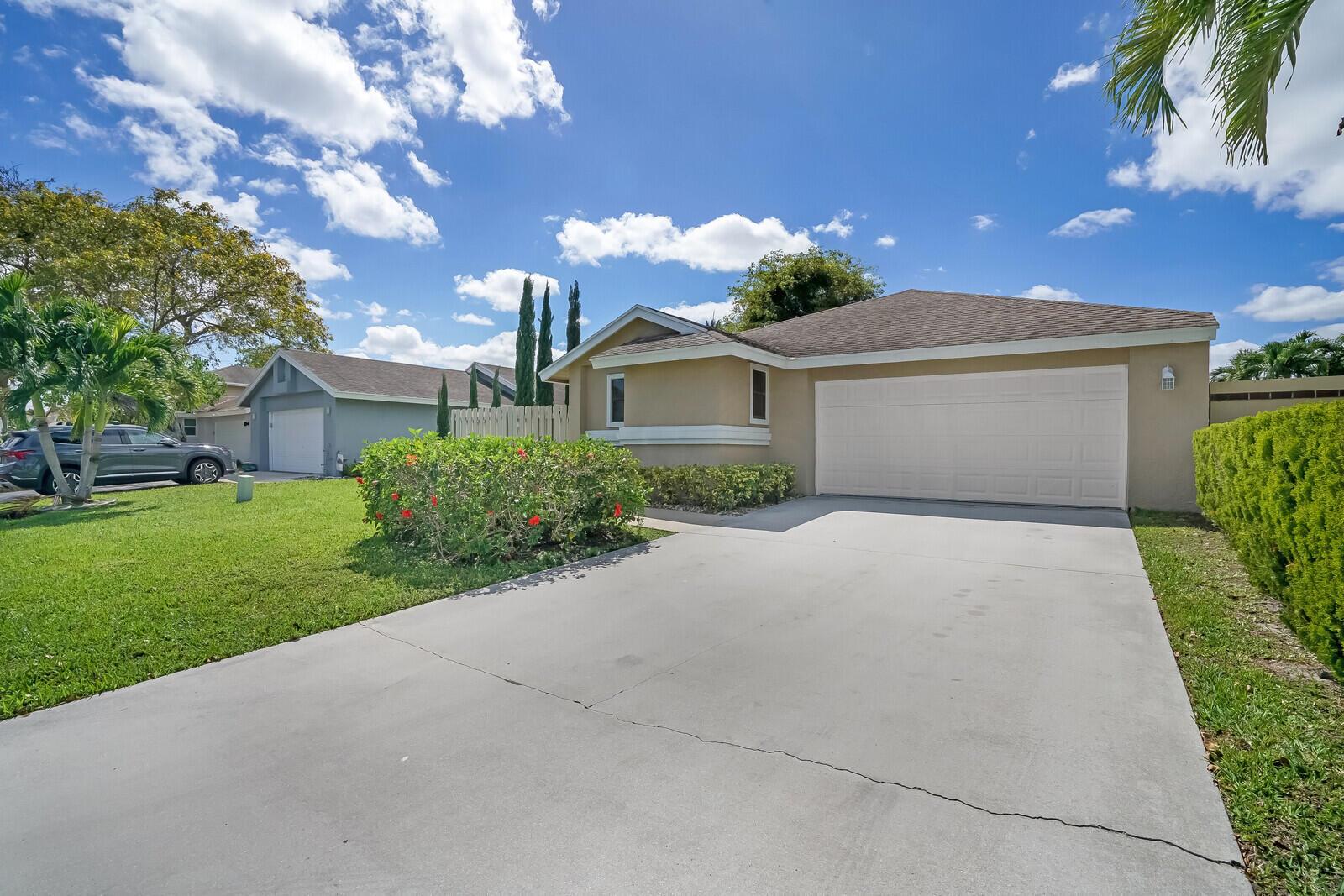 A tan single-story house with a two-car garage and a concrete driveway under blue skies — 1080 NW 19th Terrace, Delray Beach FL 33445, DELRAY BEACH, FL
