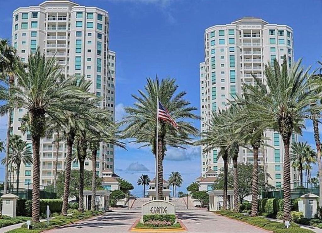 Two tall residential towers stand on either side of a palm-lined entrance with a flag — 1180 GULF BOULEVARD UNIT 606, CLEARWATER BEACH, FL