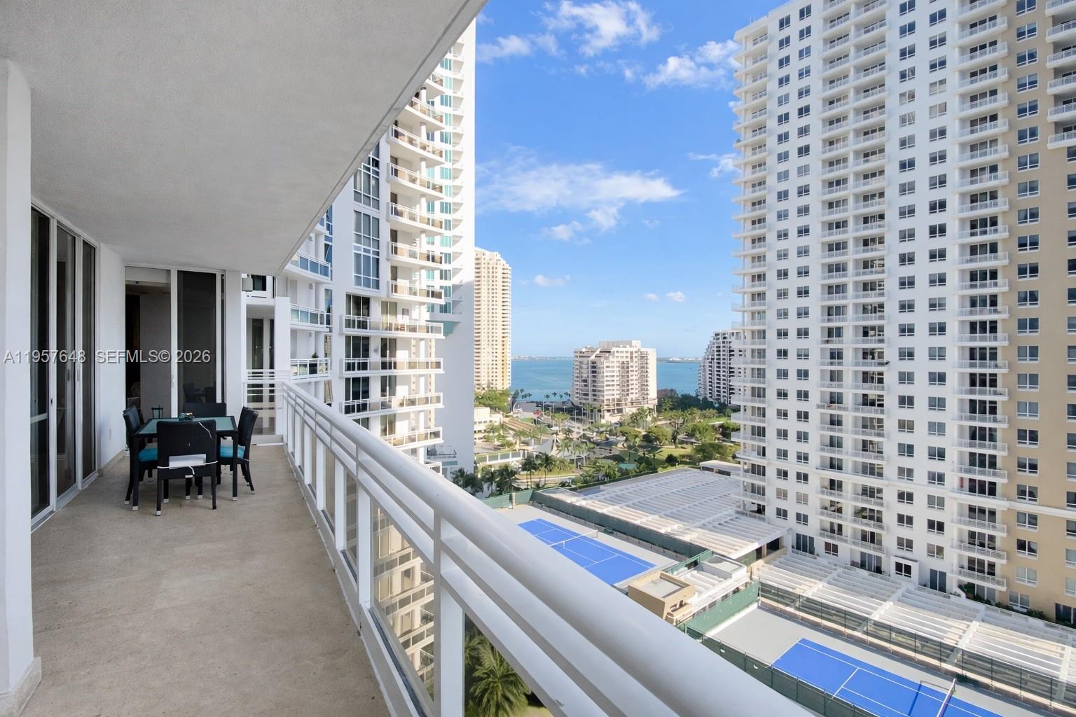 A balcony with a dining set overlooks high-rise buildings, tennis courts, and the ocean — 901 Brickell Key Blvd # 1603, Miami FL 33131, MIAMI, FL