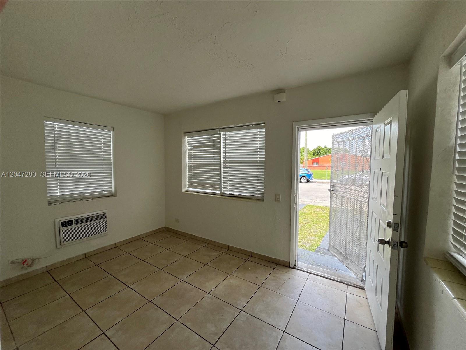 An empty room features tiled floors, white walls, window blinds, a wall-mounted air conditioner, and an open door — 733 NW 4th Ave, Miami FL 33136, MIAMI, FL